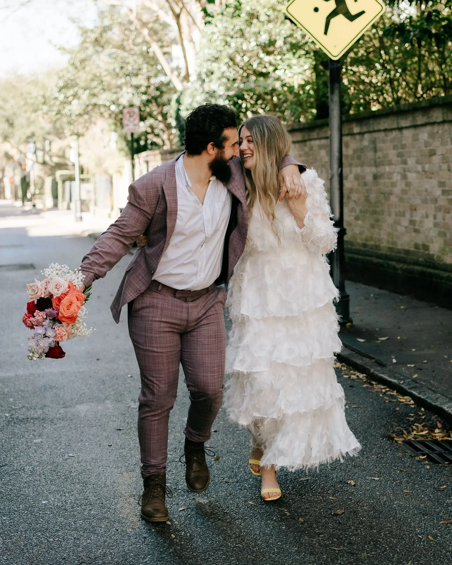 Bride and groom laughing as they walk down a leafy city street, bride holding a bouquet of pink and red flowers