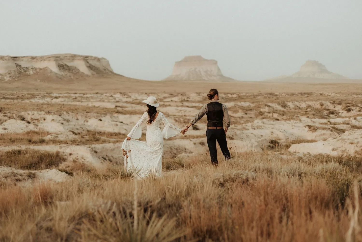 Couple in wedding clothes walking hand in hand across a desert plain toward distant mesas